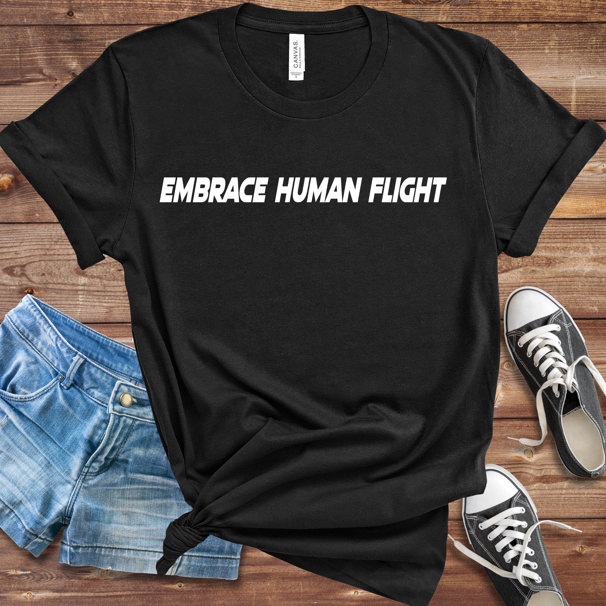 Black t-shirt with 'EMBRACE HUMAN FLIGHT' text on a wooden surface with shorts and sneakers.